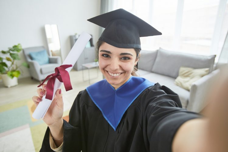 Smiling young woman wearing graduation gown while taking selfie at home or video blogging