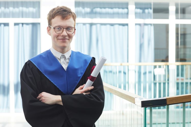 Waist up portrait of red haired young man wearing graduation gown and holding diploma while standing with arms crossed in modern university interior and smiling at camera, copy space